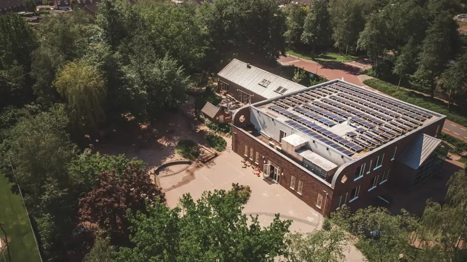 Een bakstenen gebouw met zonnepanelen op het dak, van bovenaf gezien tussen de bomen
